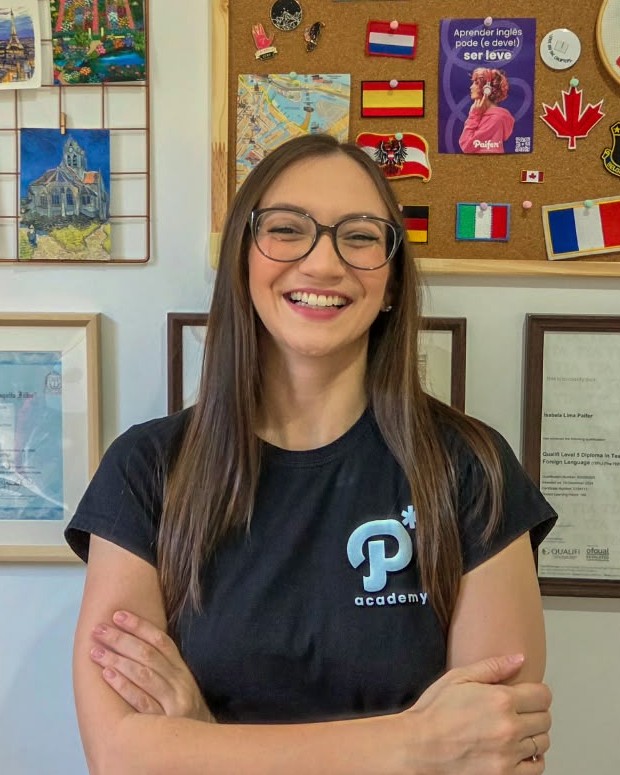 Teacher Isa sorrindo, de braços cruzados, em frente ao mural de bandeiras e aos diplomas